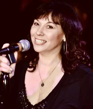 Female singer performing on stage, smiling at the microphone with hoop earrings and a black outfit.
