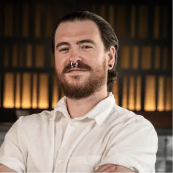 Portrait of a bearded man with a nose ring and cross-armed pose, wearing a white shirt, smiling slightly against a warm, shelves backdrop.