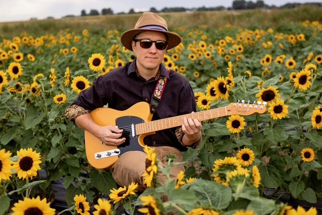 Man wearing a straw hat and sunglasses, playing a yellow electric guitar in a field of tall sunflowers.